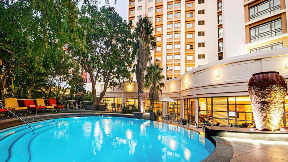 Et opplyst utendørsbasseng omgitt av grønne planter og en bygning i bakgrunnen. (AI-generert tekst)