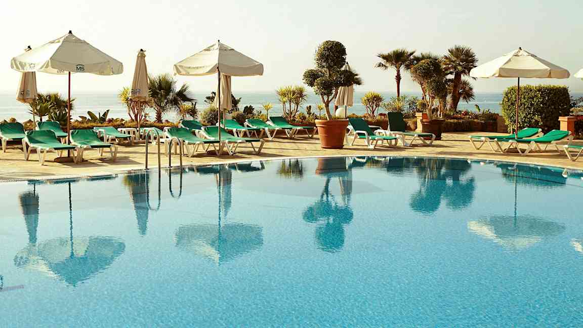 Et basseng omgitt av solsenger og parasoller, med utsikt mot havet og omkringliggende planter. (AI-generert tekst)