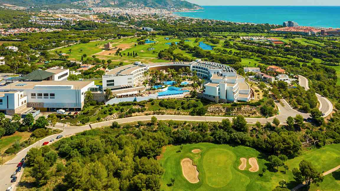 Utsikt ovenfra over et hotellområde omgitt av grøntområder og en golfbane. Havet og en by er i bakgrunnen. (AI-generert tekst)