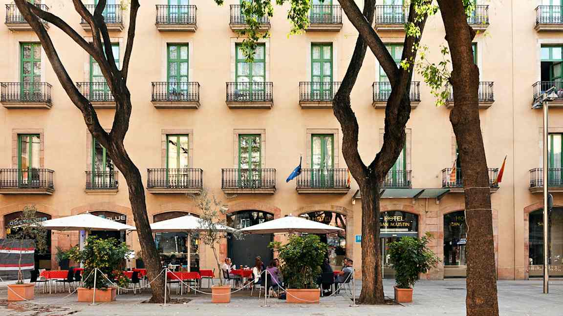 Et hotell med flere etasjer og balkonger, en fortau med parasoller og sitteplasser blant trær. (AI-generert tekst)