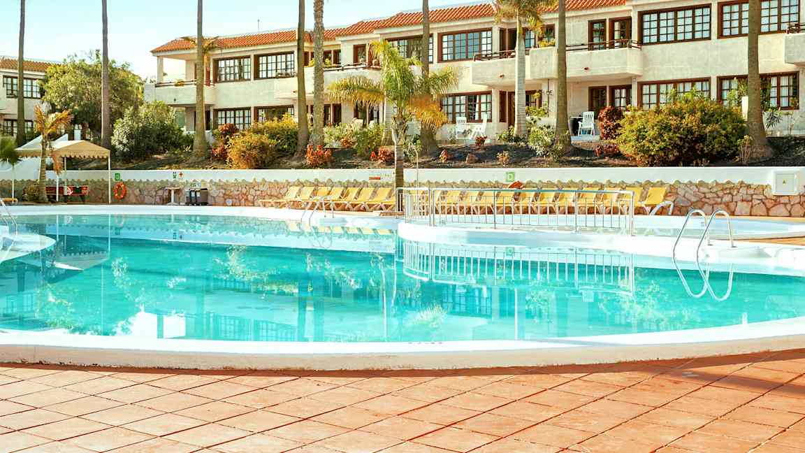 Et svømmebasseng omgitt av solsenger med en hvit bygning i bakgrunnen. (AI-generert tekst)