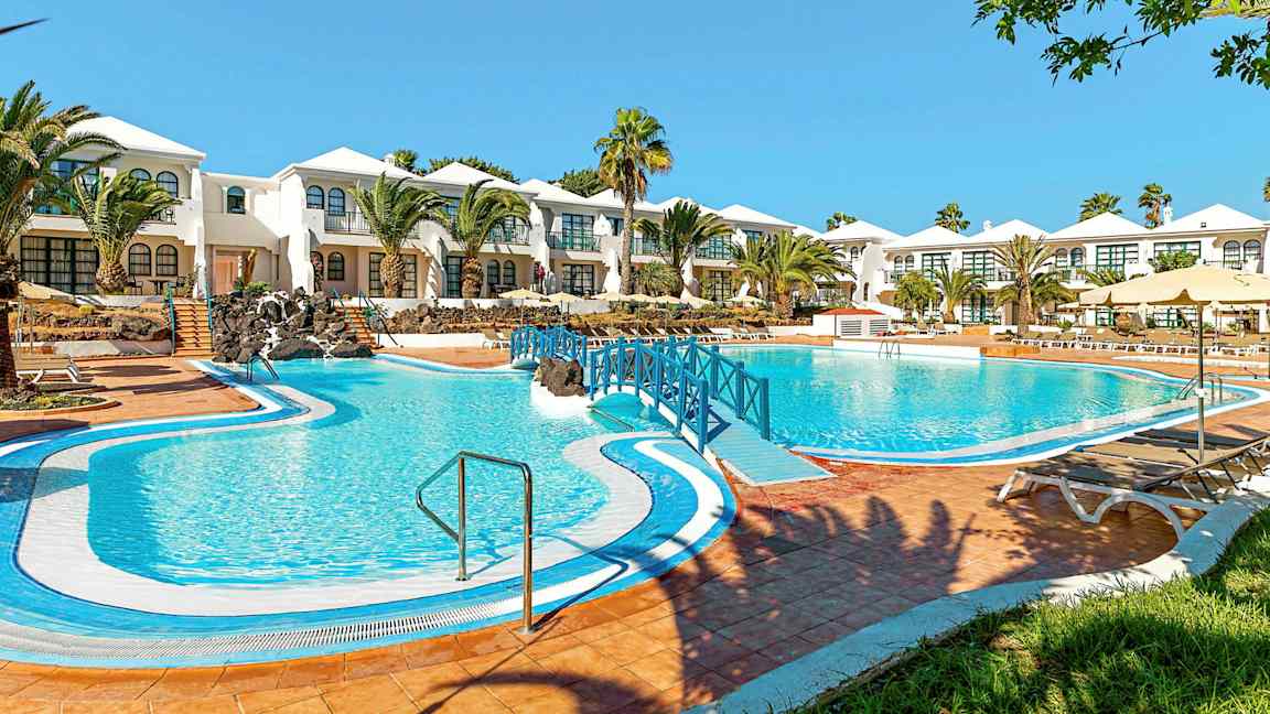 Et hotell med hvitmalte bygninger og et stort blått basseng foran, omgitt av palmer og grønne områder. (AI-generert tekst)