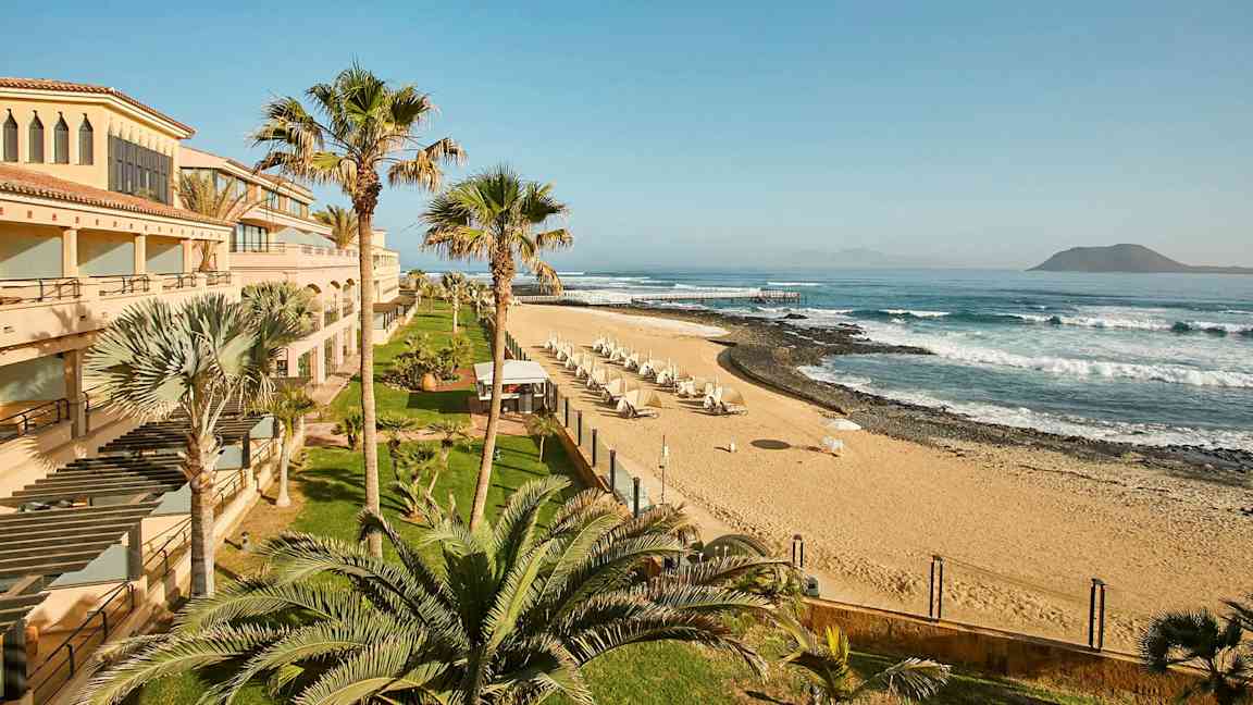 En strand med sand, parasoller og bølger, omgitt av bygninger og palmer. (AI-generert tekst)