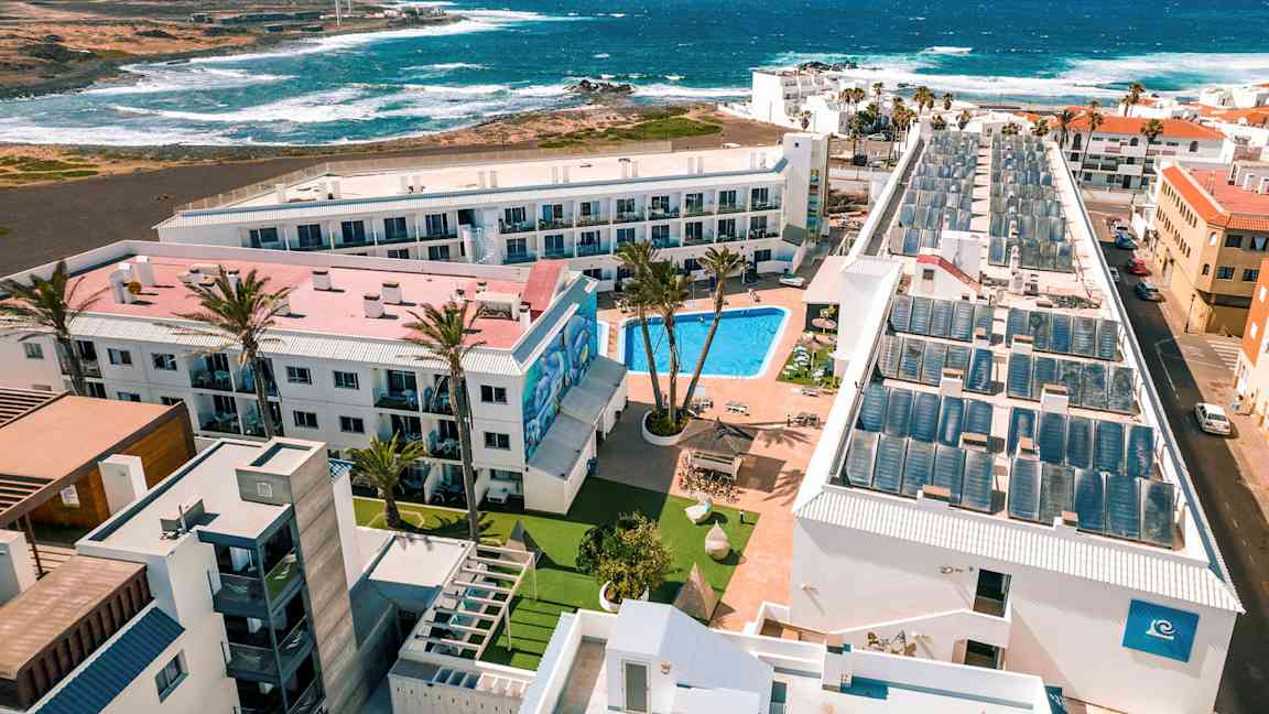 Et hotellresort med basseng, omgitt av bygninger, med utsikt til havet og bølgene. (AI-generert tekst)