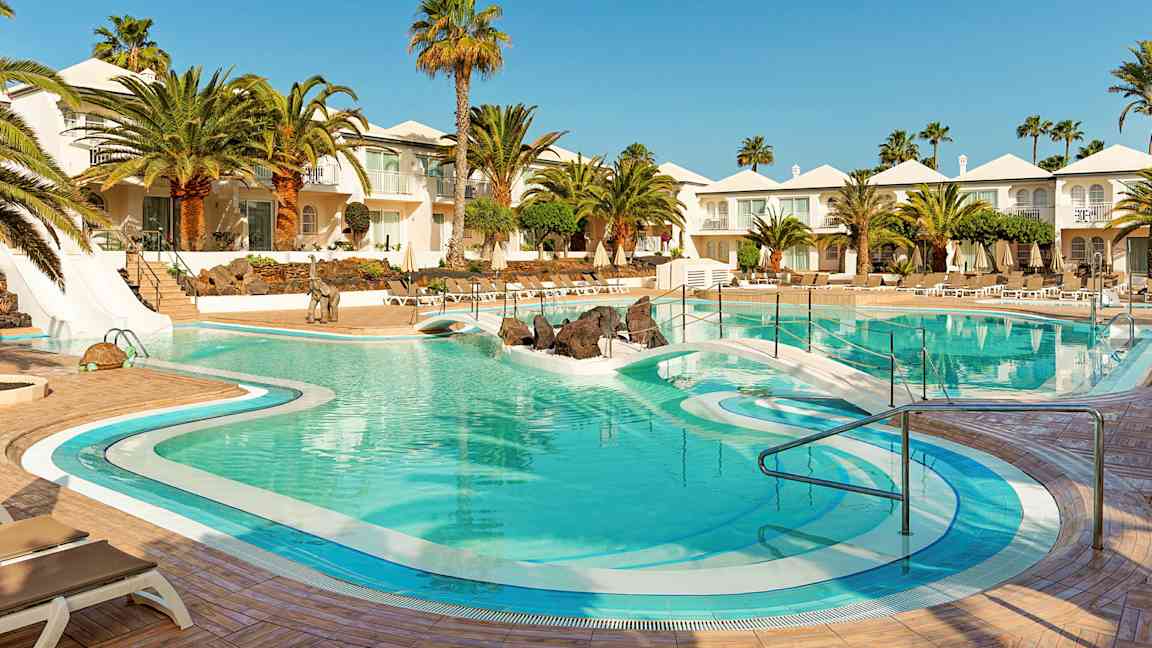 Et hotellbasseng omgitt av palmer, solsenger og hvite bygninger. (AI-generert tekst)