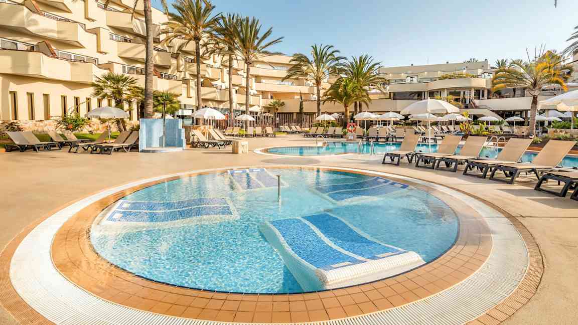 Et bassengområde på et hotell med solsenger og parasoller rundt bassenget, omgitt av palmer og balkonger. (AI-generert tekst)