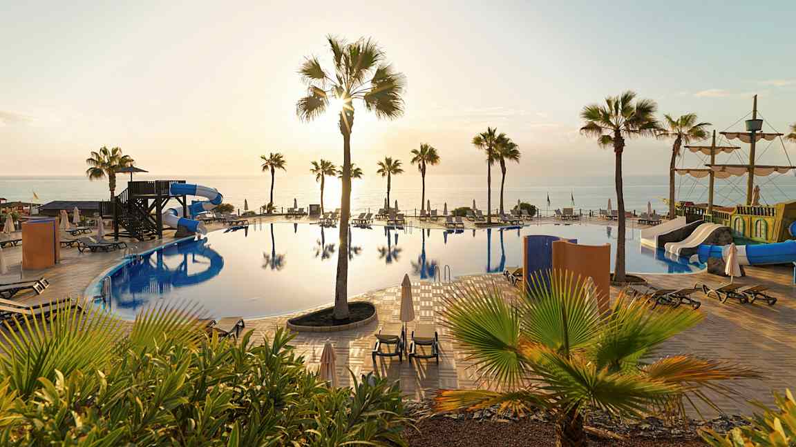 Bilde av et basseng med vannsklier, solsenger og palmer, samt havet i bakgrunnen ved solnedgang. (AI-generert tekst)