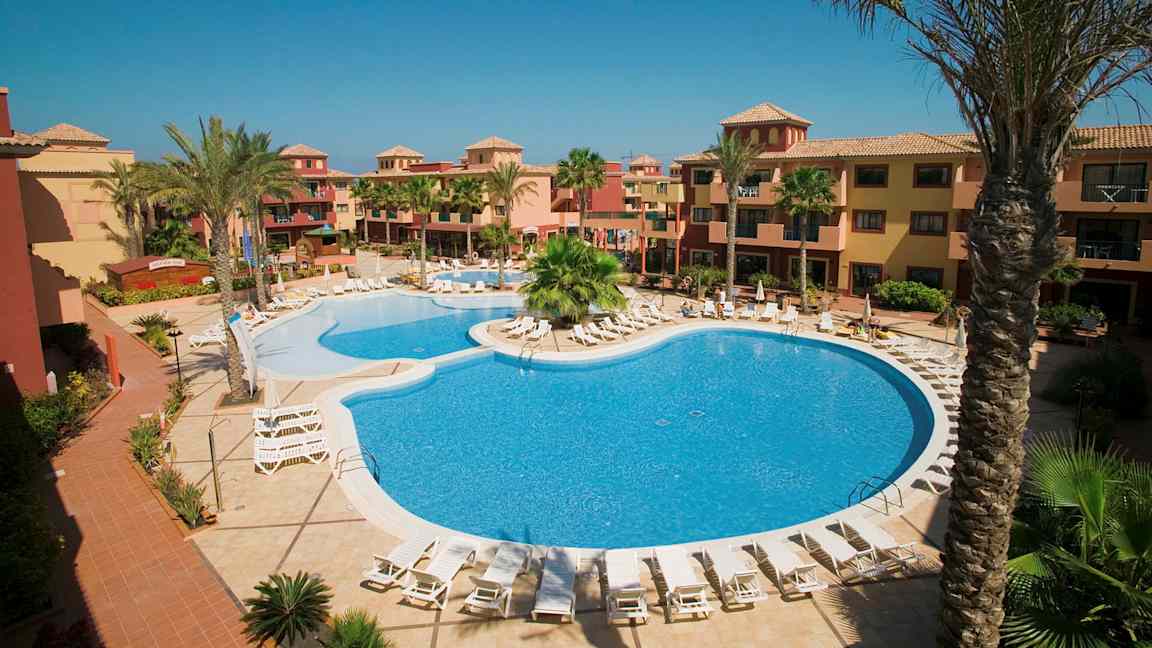 Hotellområde med basseng og solstoler, omgitt av palmer og bygninger i varme farger. (AI-generert tekst)