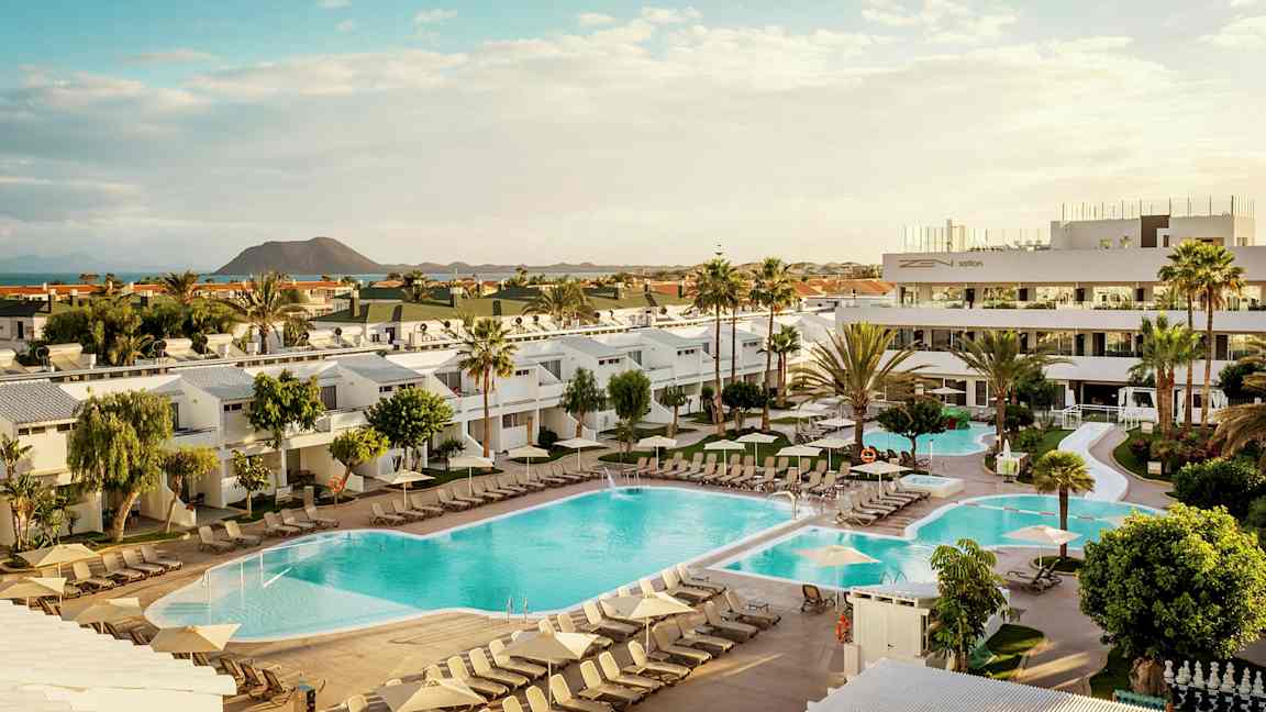 Bilde av et hotellområde med bassenger, solsenger og omkringliggende bygninger, samt en vulkan i bakgrunnen. (AI-generert tekst)