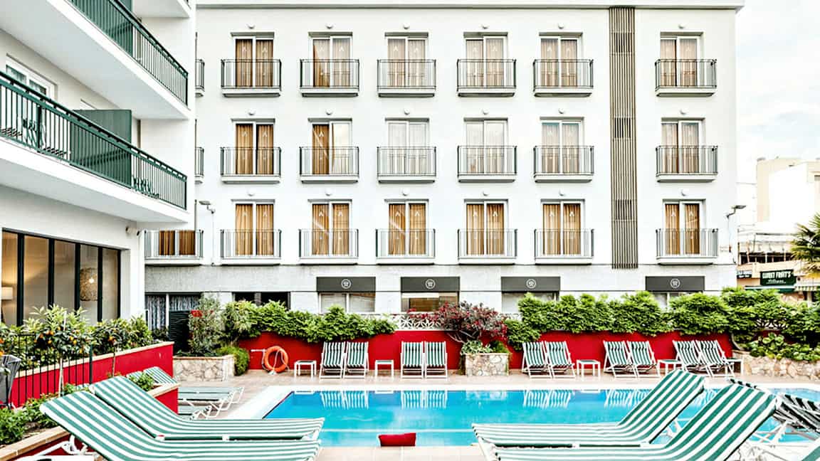 Bilde av en hotellbygning med hvit fasade og flere balkonger. Foran bygningen er et utendørsbasseng med stripete solsenger plassert rundt. (AI-generert tekst)