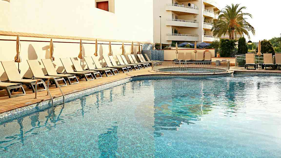 Et utendørs svømmebasseng omgitt av solsenger og parasoller, med en bygning og en palme i bakgrunnen. (AI-generert tekst)