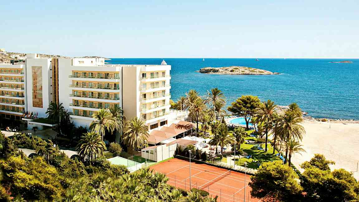 Bilde av et feriested ved stranden med en bygning, palmer, basseng og tennisbaner, lokalisert ved sjøen. (AI-generert tekst)