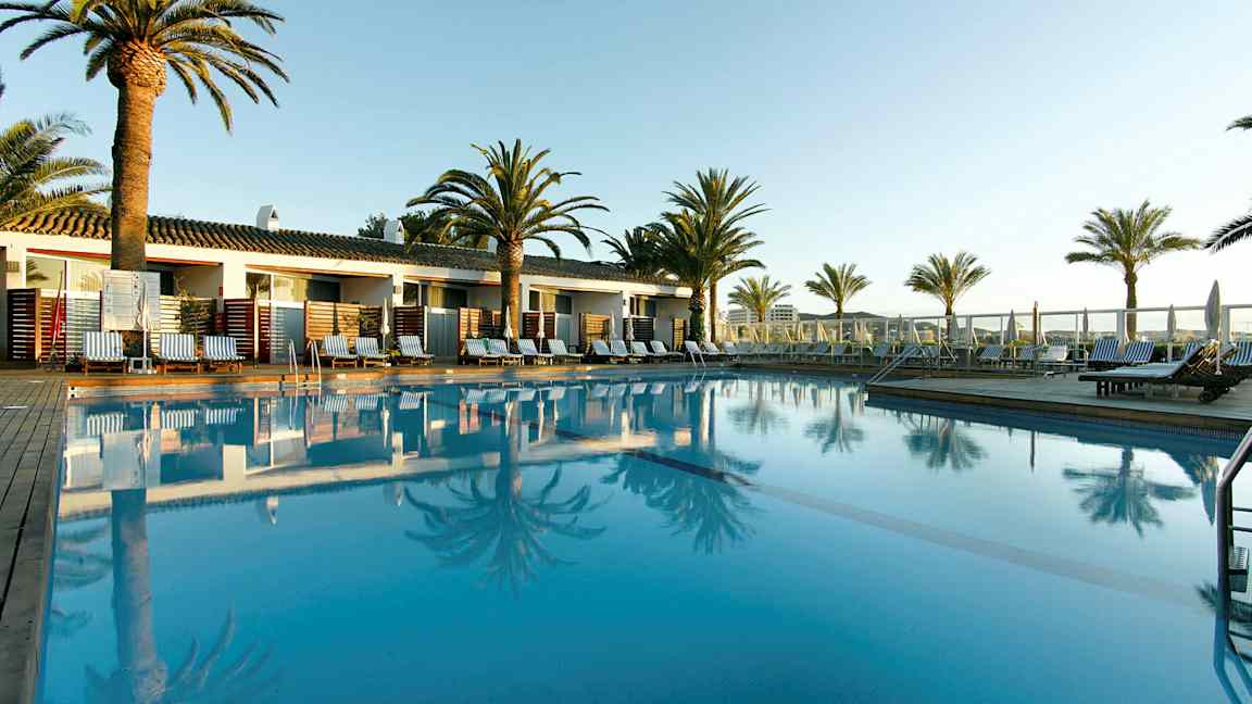 Et utendørs basseng omgitt av solsenger, palmer og bygninger i bakgrunnen. (AI-generert tekst)