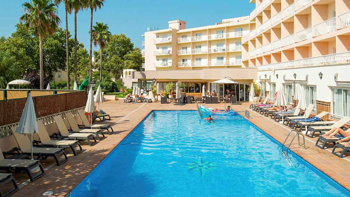Et hotellbygg ligger ved siden av et utendørsbasseng med solsenger og parasoller rundt. (AI-generert tekst)