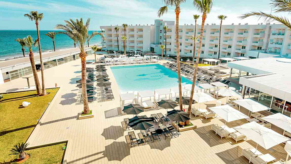 En utsikt over et feriested med et stort basseng, omgitt av solsenger og parasoller, med palmer og en bygning i bakgrunnen nær havet. (AI-generert tekst)