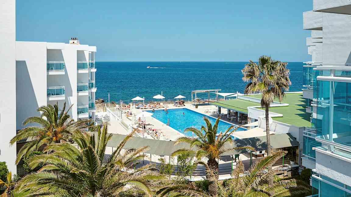 Et bilde av et hotellområde med utsikt over havet. Det er et svømmebasseng omgitt av solsenger og parasoller, omgitt av palmer og hvite bygninger. (AI-generert tekst)