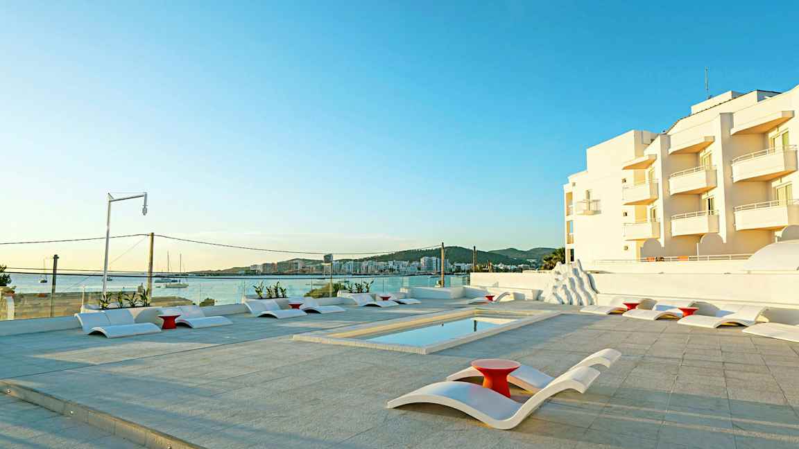 Utendørs bassengområde med moderne solsenger og utsikt over sjøen og hvite bygninger i bakgrunnen. (AI-generert tekst)