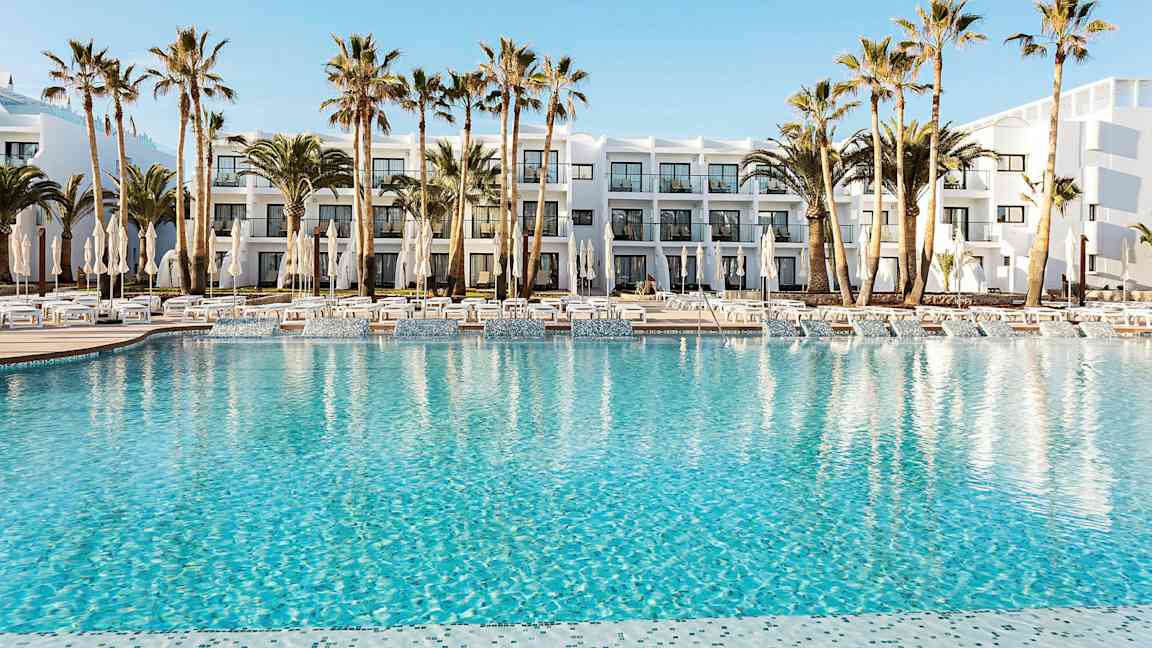 Hotellets bassengområde med klart blått vann, omgitt av solsenger, parasoller og palmer. I bakgrunnen sees en hvit bygning med balkonger. (AI-generert tekst)