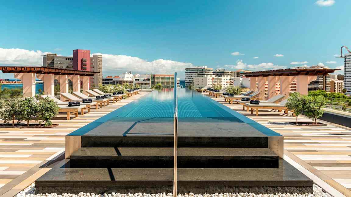 Et basseng på taket med solsenger og utsikt over byen under blå himmel. (AI-generert tekst)