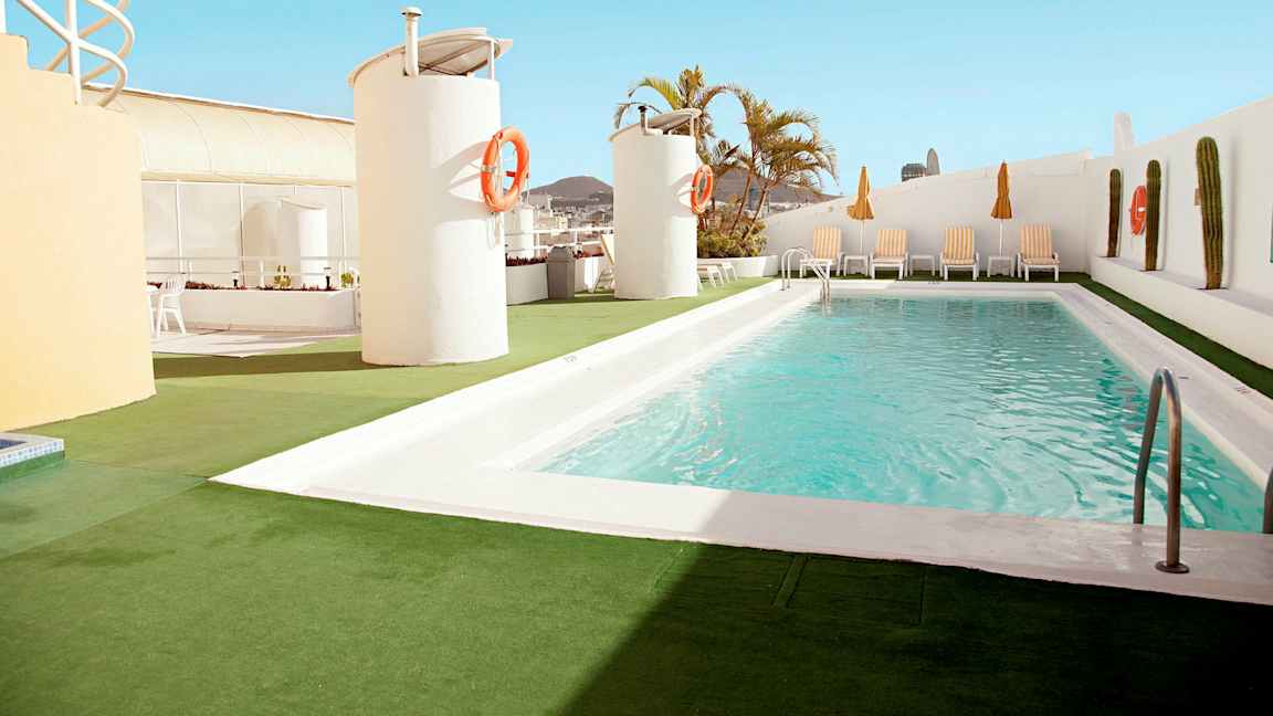 Et utendørs basseng ligger på en takterrasse med solsenger og parasoller rundt. (AI-generert tekst)