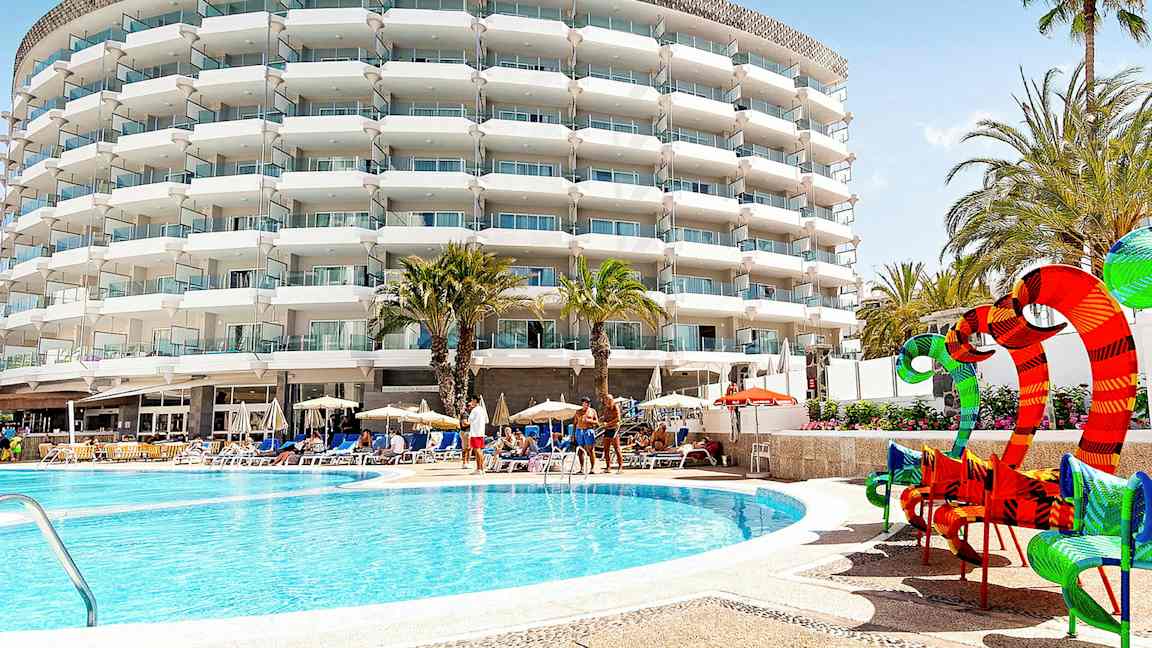 Hotellbygning med balkonger, palmer og bassengområde. (AI-generert tekst)