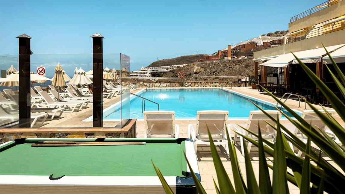 Utendørs svømmebasseng med solsenger og parasoller rundt, med utsikt mot bygninger i bakgrunnen på en skråning. (AI-generert tekst)