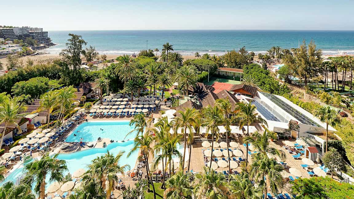 Utsikt over et feriekompleks med bassenger, solsenger og palmer nær en sandstrand ved havet. (AI-generert tekst)