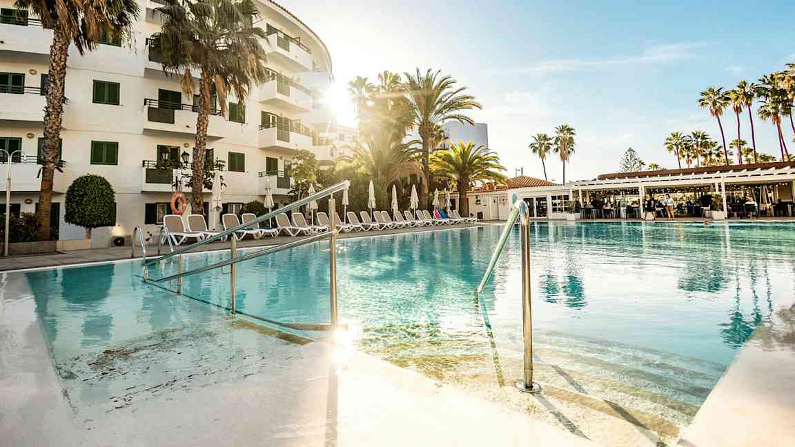 Et utendørs basseng foran et hotell omgitt av palmer og solsenger. (AI-generert tekst)