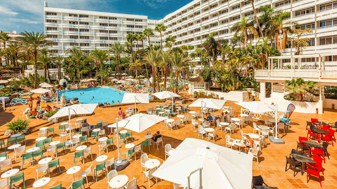 Hotell med stor utendørsterrasse, bassengområde og palmer. (AI-generert tekst)