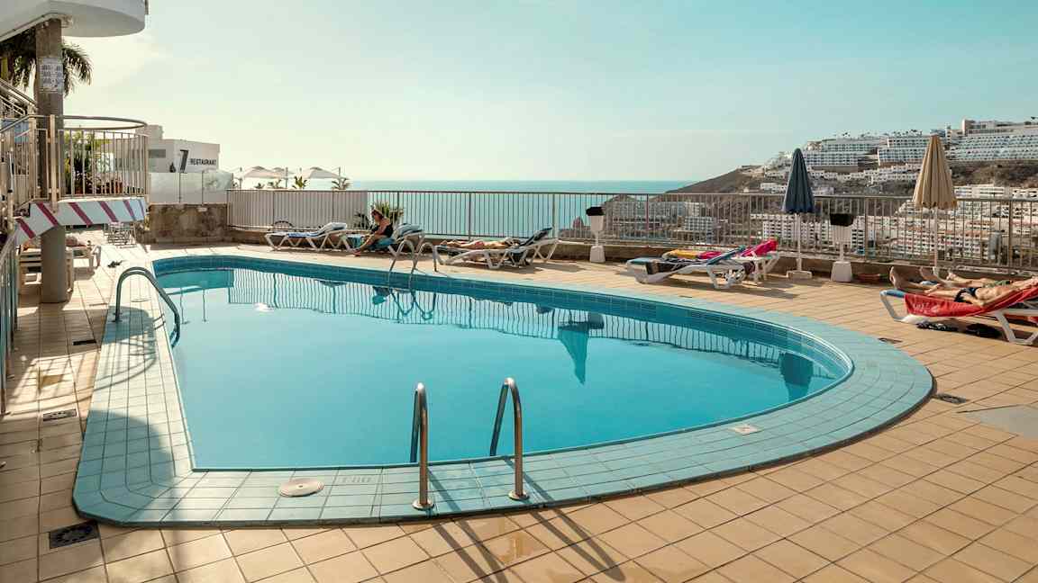 Et utendørs svømmebasseng med solsenger og parasoller rundt, med utsikt over havet og omkringliggende bygninger i bakgrunnen. (AI-generert tekst)