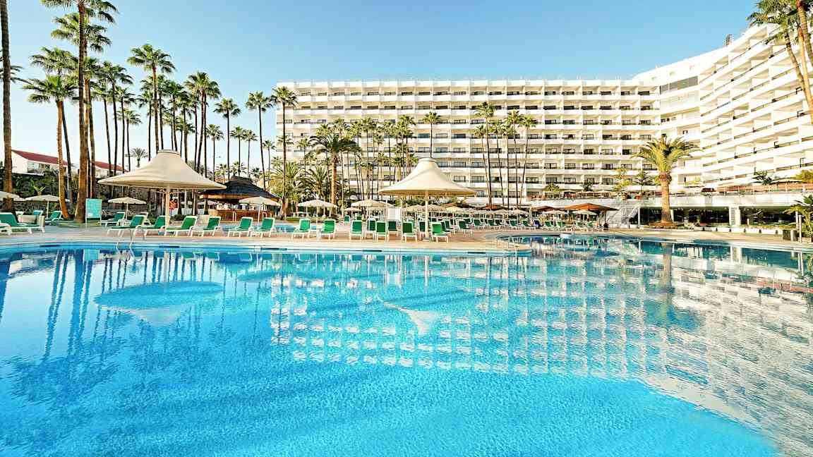 Hotellbygning med stort basseng og solsenger i forgrunnen, omgitt av palmer. (AI-generert tekst)