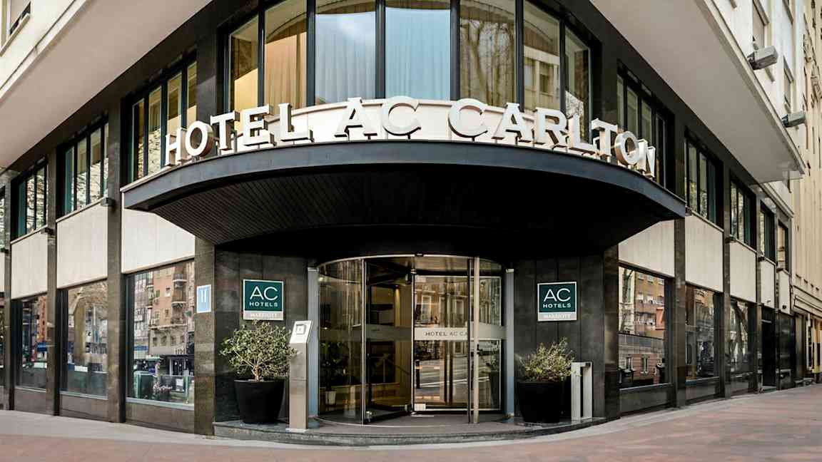 En hotellfasade med skilteteksten "HOTEL AC CARLTON" synlig over inngangen. (AI-generert tekst)