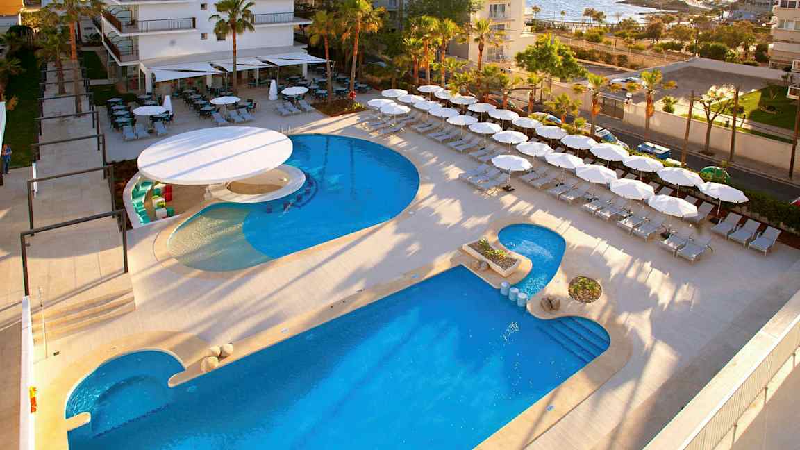 Hotellområde nær sjøen med basseng, solsenger og parasoller. (AI-generert tekst)
