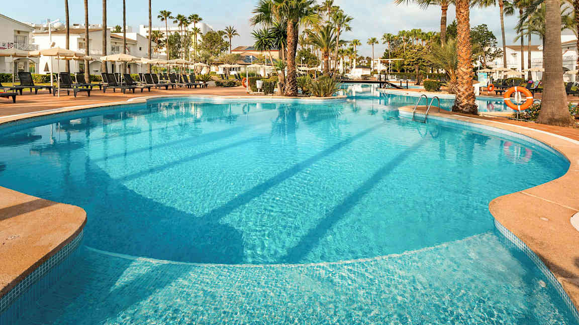 Stor utendørs basseng med solsenger og parasoller omgitt av palmer og bygninger. (AI-generert tekst)