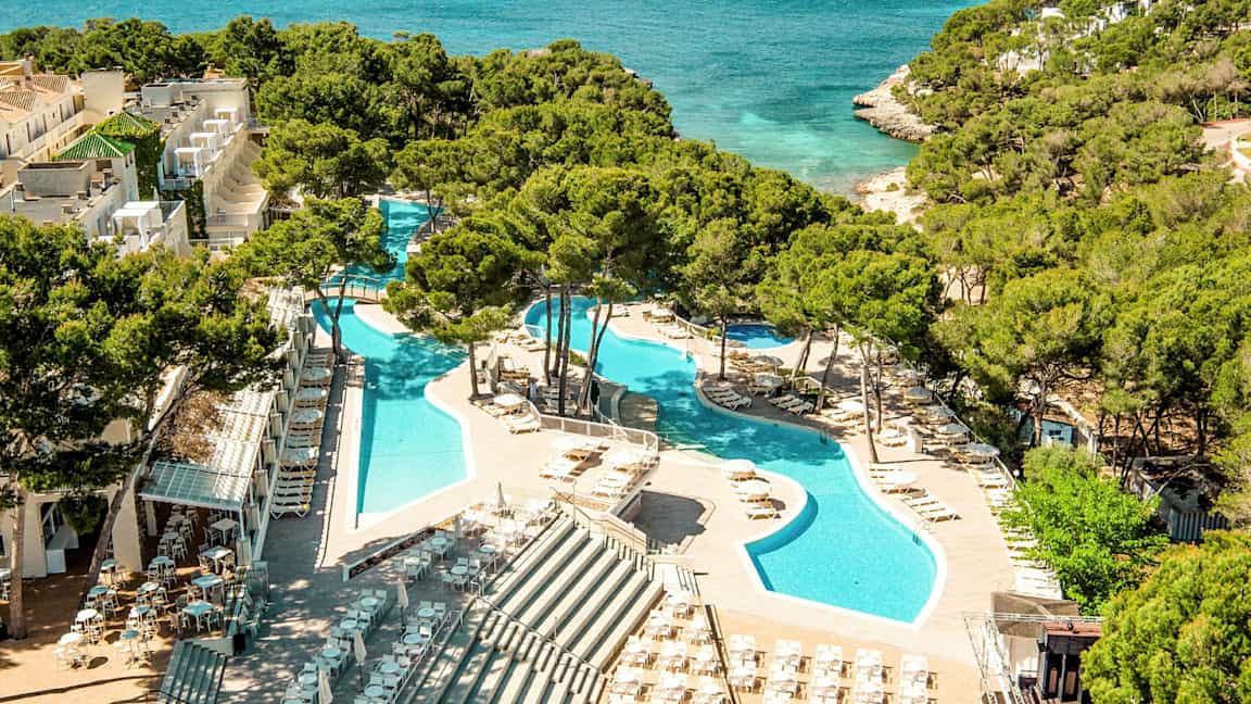 En utsikt over et feriested med bassenger omgitt av solsenger og parasoller, plassert ved siden av et turkist hav med frodig vegetasjon i bakgrunnen. (AI-generert tekst)