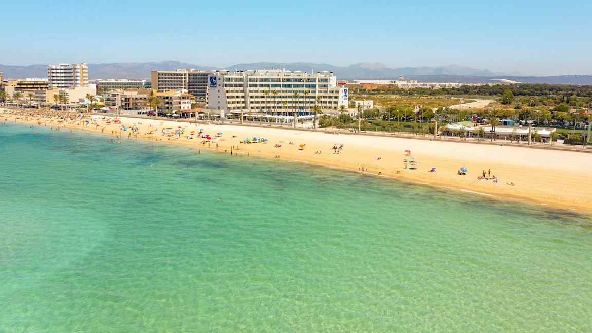 Sandstrand med badende personer og klart turkisblått vann. Hoteller og bygninger i bakgrunnen. (AI-generert tekst)