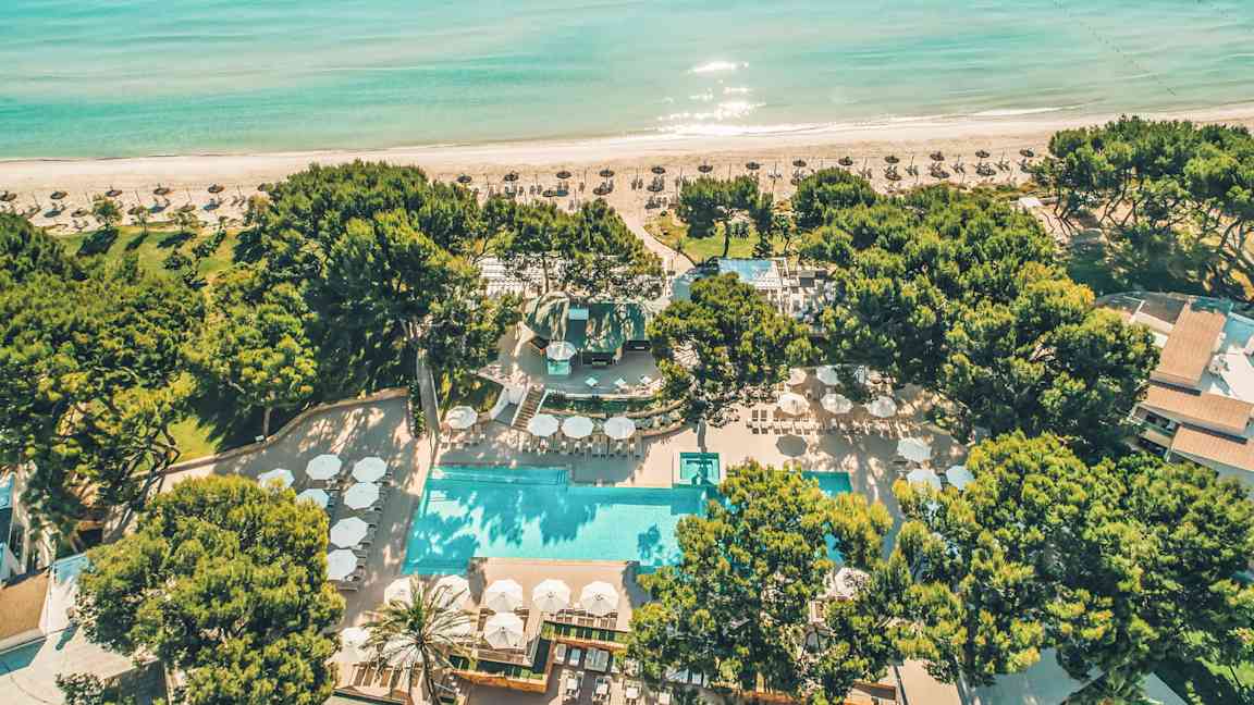 Flyfoto av en strandresort med basseng, omgitt av trær og stranden i bakgrunnen. (AI-generert tekst)