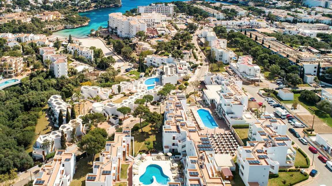 Luftfoto av et kystnært ferieområde med hvite bygninger og svømmebassenger. (AI-generert tekst)
