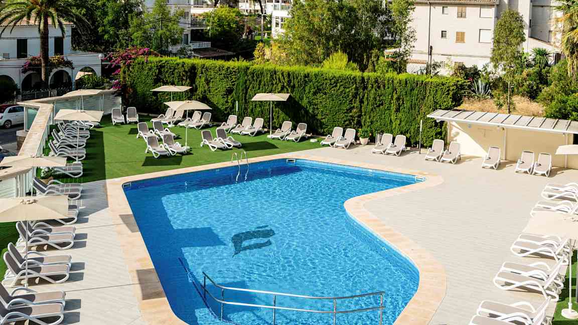 Utendørsbasseng med solsenger og parasoller ved siden, omgitt av grøntområder og bygninger. (AI-generert tekst)