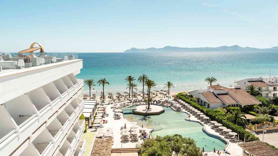 En utsikt over hotellbasseng, strand og hav med palmer og bygninger. (AI-generert tekst)