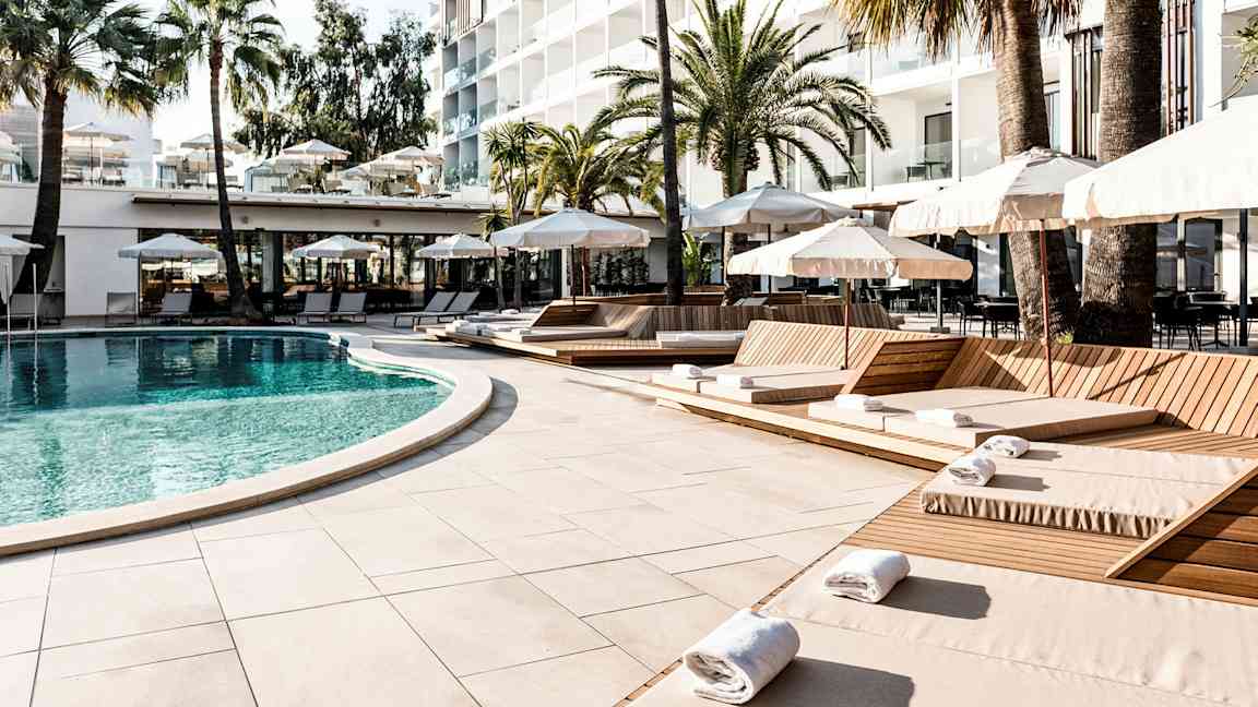 Et basseng omgitt av solsenger og parasoller ved et hotell, med palmer i bakgrunnen. (AI-generert tekst)