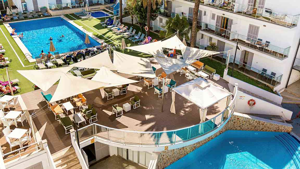 Et bilde av et hotellkompleks med et stort basseng omgitt av solsenger og parasoller. I forgrunnen er en utendørsbar med mange sitteplasser under skyggefulle seil. (AI-generert tekst)
