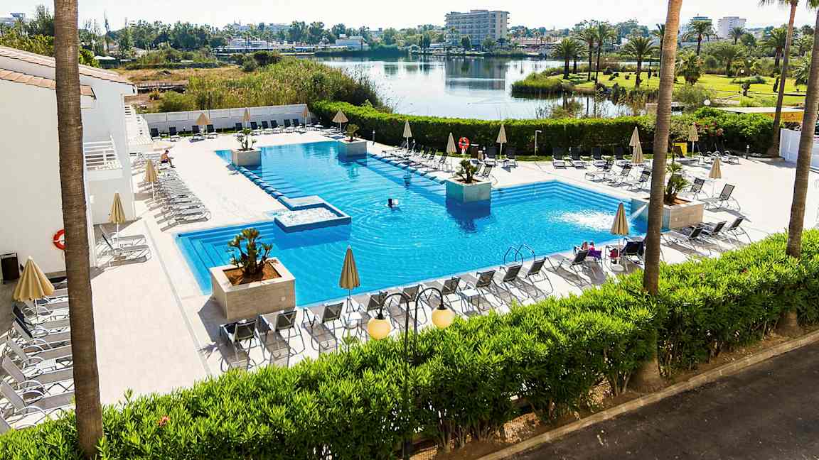 Utendørs svømmebasseng med solsenger og parasoller rundt, omgitt av grønne planter og med utsikt over en innsjø og et åpent landskap. (AI-generert tekst)