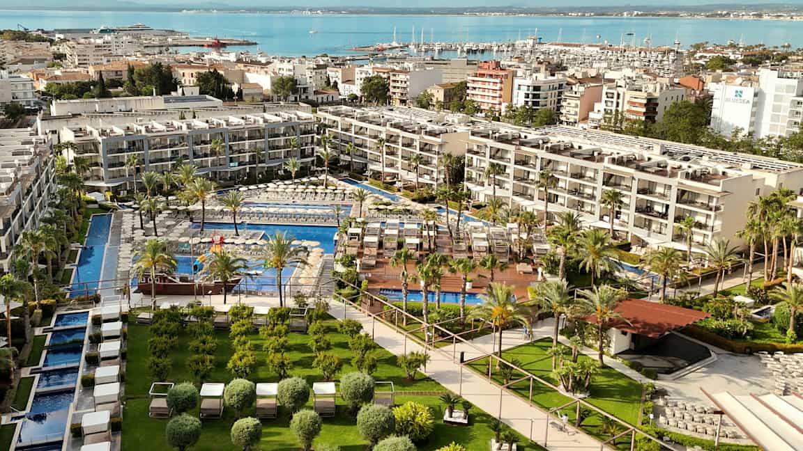 Et hotellkompleks med bassenger omgitt av grøntområder og solsenger, plassert ved vannet med en havn i bakgrunnen. (AI-generert tekst)