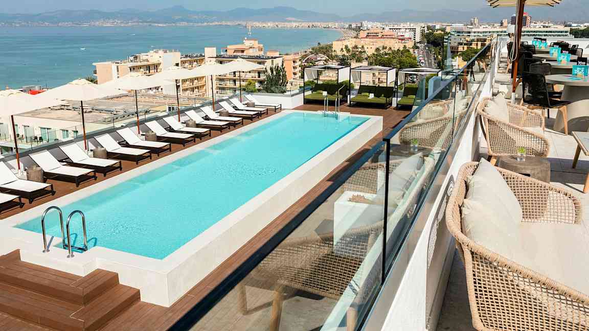 Takleilighet med et utendørsbasseng omgitt av solsenger og skygge, med utsikt over en kystby. (AI-generert tekst)