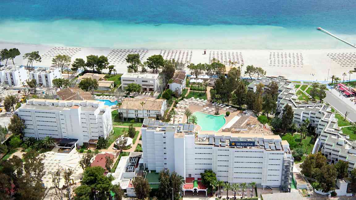 Luftfoto av et hotellområde nær stranden med parasoller på sanden og en klarblå sjøutsikt. (AI-generert tekst)