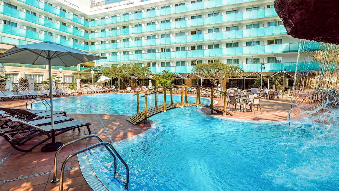 Et utendørs svømmebasseng ved en hotellbygning med solsenger, bord og parasoller. En liten bro over bassenget og en del av en fossestruktur er synlig. (AI-generert tekst)