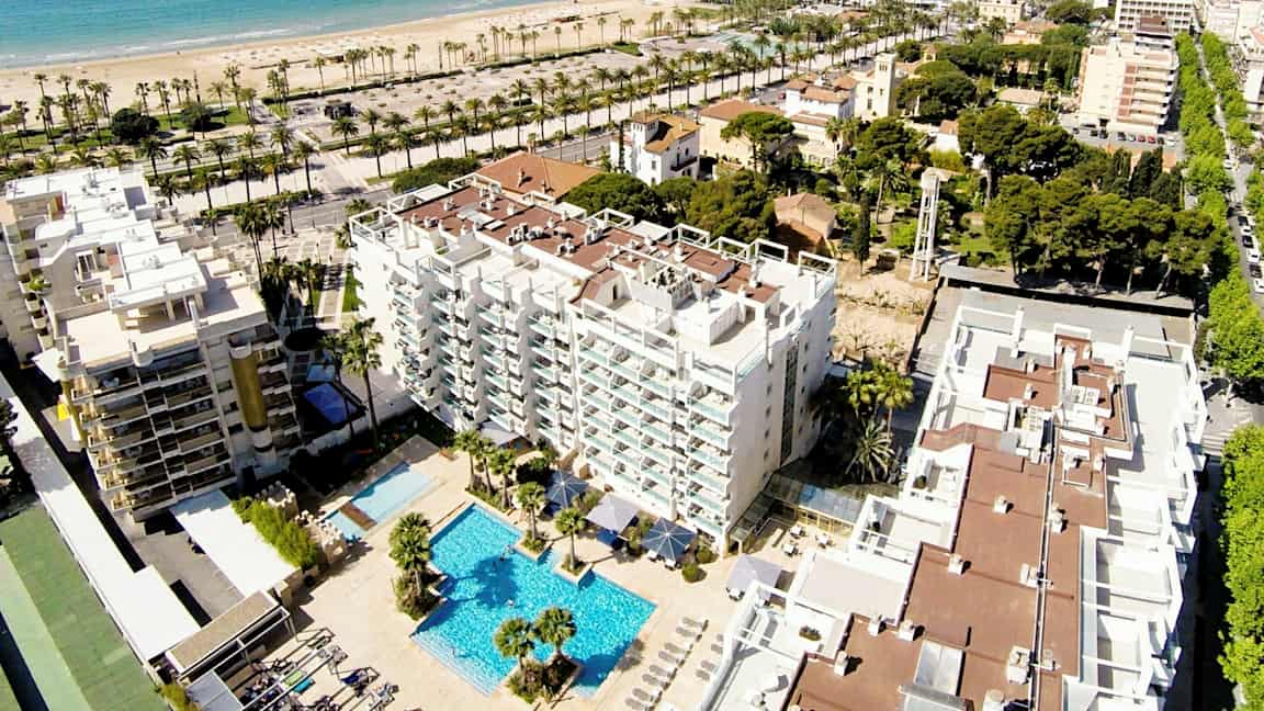Luftfoto av et hotell med bassengområde, omgitt av bygninger og grøntarealer. I bakgrunnen ses en sandstrand og havet. (AI-generert tekst)