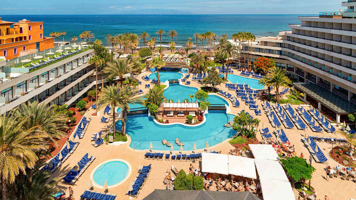 Det er en utsikt over et hotellområde med flere bassenger, omgitt av solsenger og parasoller, samt trær og bygninger. I bakgrunnen er havet synlig. (AI-generert tekst)