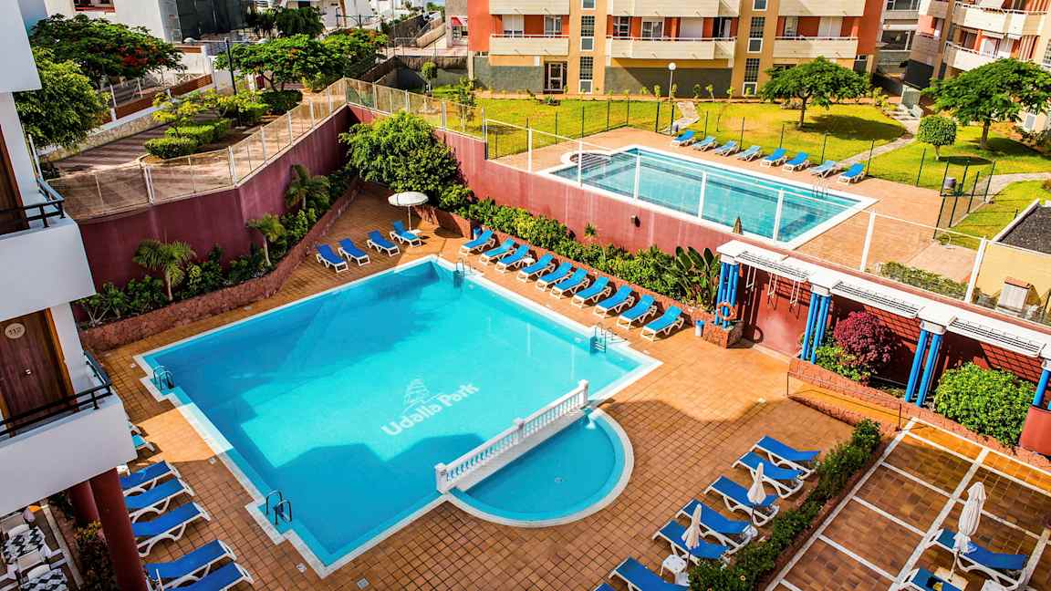 Et hotellområde med to bassenger, solsenger og omkringliggende bygninger. (AI-generert tekst)